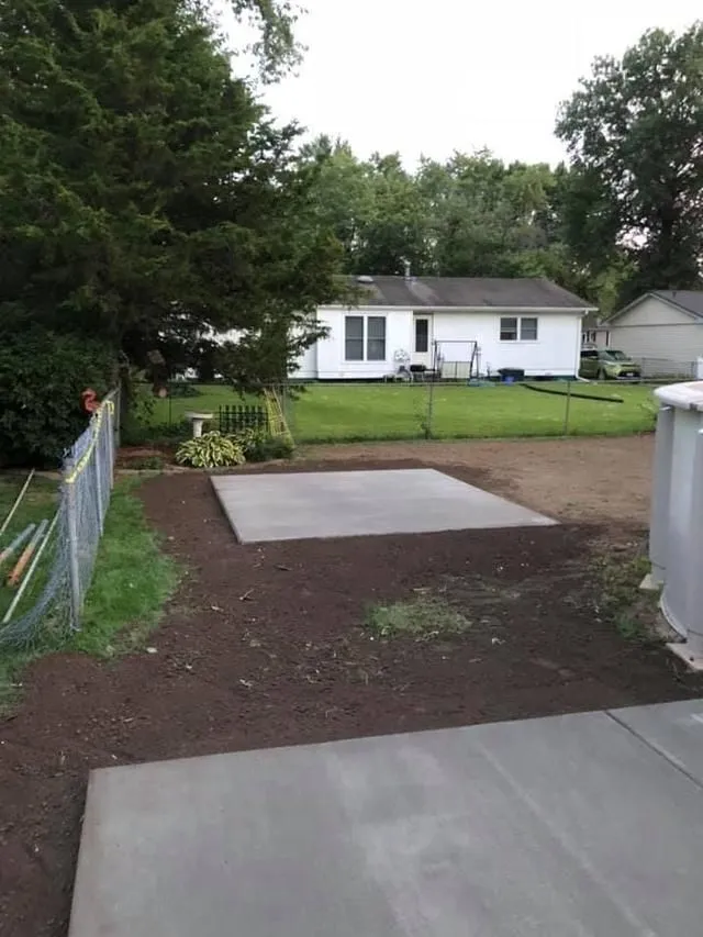 A concrete slab in a backyard with a house, chain-link fence, and trees.