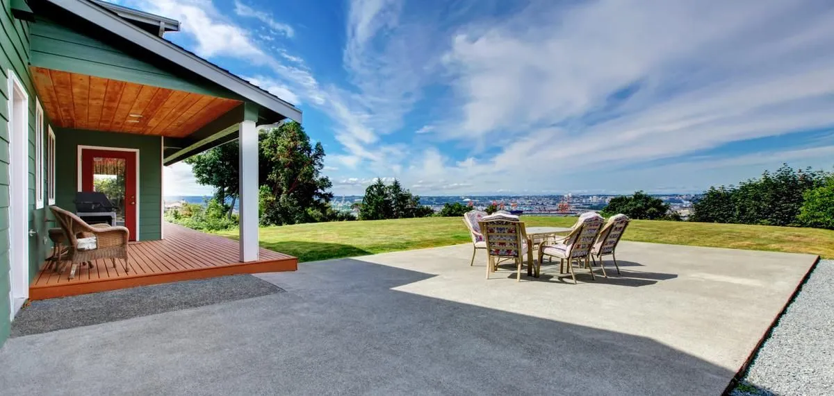 Exterior view of a house with a porch and concrete patio, with a lawn and distant cityscape under a blue sky.
